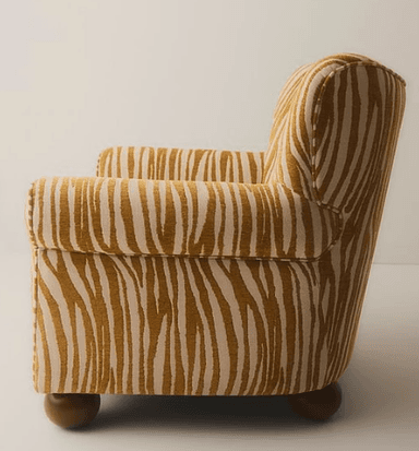 A compact upholstered armchair featuring a distinctive gold and off-white zebra-print fabric. It has a high, subtly winged backrest and classic rolled armrests. The chair is supported by four short, dark wooden bun feet, and shows visible piping detail along its seams. — 3D model