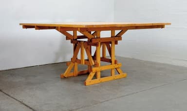 A wooden dining table with a rectangular, dual-sectioned tabletop and a distinct trestle-style base. The tabletop appears to be constructed from planks and shows a visible seam down the center, suggesting it might be extendable or foldable. The base is composed of angled wooden beams forming a robust, scaffold-like structure with cross-bracing for stability. The overall design has a rustic or industrial aesthetic. — 3D model