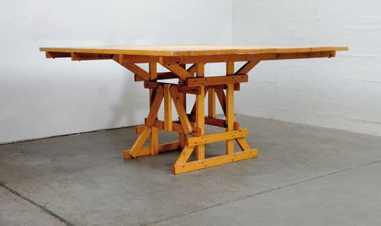 A wooden dining table with a rectangular, dual-sectioned tabletop and a distinct trestle-style base. The tabletop appears to be constructed from planks and shows a visible seam down the center, suggesting it might be extendable or foldable. The base is composed of angled wooden beams forming a robust, scaffold-like structure with cross-bracing for stability. The overall design has a rustic or industrial aesthetic. — 3D model