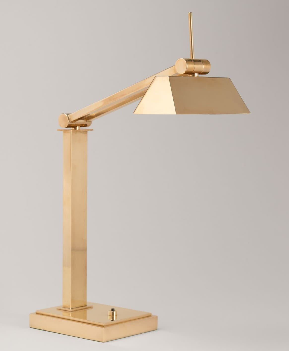 A sleek, modern desk lamp crafted from polished brass, featuring an adjustable arm and a geometric, trapezoidal shade. The lamp stands on a rectangular base with an integrated toggle switch. — 3D model