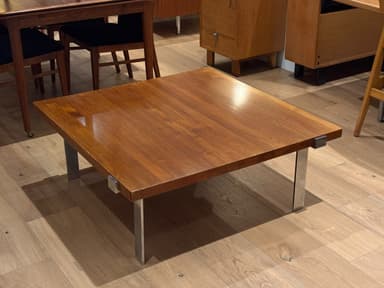 A square coffee table featuring a wooden top and four recessed metal legs. The tabletop has a polished, reddish-brown finish, suggesting a teak or walnut construction. The legs are made of polished chrome or stainless steel, with a rectangular profile, and are integrated into the corners of the table, giving it a minimalist and sleek appearance. The table exhibits characteristics of Mid-Century Modern design. — 3D model