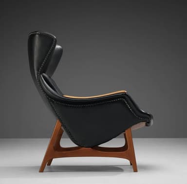 A high-backed armchair with a distinctive, sculptural form. The seat and backrest are upholstered in black material, possibly leather, and accented with numerous small decorative studs along the edges. The arms appear to be crafted from a lighter-colored wood, contrasting with the main upholstery. The chair is supported by a sculpted wooden base with a dark wood finish, featuring angled legs that provide stability. — 3D model