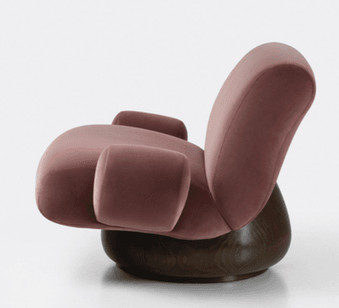 A plush armchair with a rounded silhouette and voluminous cushioning. The seat and backrest are upholstered in a soft, rose-colored velvet fabric. It features integrated armrests that flow seamlessly from the main body of the chair. The chair is supported by a substantial, dark-stained wooden base with a rounded, drum-like shape. — 3D model