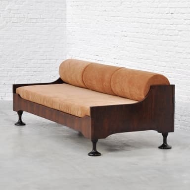 An image showing a modern daybed with a dark wood frame and light orange fabric upholstery. It features a long, rectangular seat cushion and a cylindrical bolster cushion along one side, acting as a backrest. The daybed rests on four short, dark, tapered legs. — 3D model