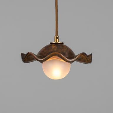 A pendant light featuring a distinctive wavy, shell-like metal shade with a mottled, patinated bronze finish. Suspended beneath the shade is a spherical frosted glass diffuser, emitting a soft, warm light. The fixture hangs from a braided textile cord, connecting to the shade via a small brass fitting. — 3D model