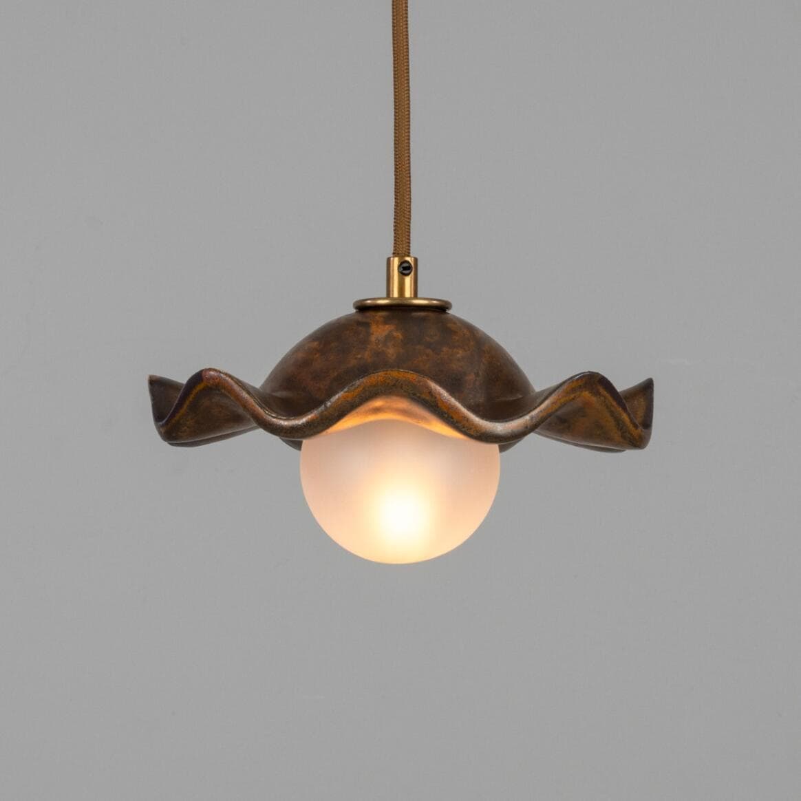 A pendant light featuring a distinctive wavy, shell-like metal shade with a mottled, patinated bronze finish. Suspended beneath the shade is a spherical frosted glass diffuser, emitting a soft, warm light. The fixture hangs from a braided textile cord, connecting to the shade via a small brass fitting. — 3D model