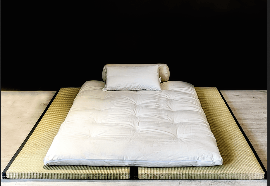 A traditional Japanese futon mattress set, comprising a white, tufted futon mattress, a rectangular pillow, and a cylindrical bolster, all placed atop two woven tatami mats. The tatami mats have black fabric borders and form a low-profile platform for the bedding. The entire arrangement is presented against a white background, highlighting its minimalist design. — 3D model