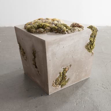 A cubic concrete ottoman with an irregular, textured top surface composed of braided and knotted yarn in shades of yellow, green, and grey. The concrete body features several irregular voids and openings, some of which reveal the same yarn material embedded within. — 3D model