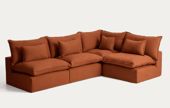A contemporary modular sectional sofa upholstered in a burnt orange fabric. It consists of multiple deep-seated sections configured into an L-shape, featuring plush back cushions and several rectangular throw pillows, all contributing to a relaxed and inviting aesthetic. The base is also fully upholstered, maintaining a consistent, clean profile. — 3D model
