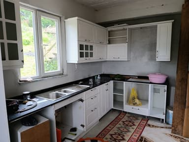 An L-shaped modular kitchen cabinet system, presented in an aerial view. It features white shaker-style cabinet doors and drawers for both upper and lower units. The lower cabinetry includes an integrated double-basin stainless steel sink within a black speckled countertop. One section of the upper cabinets features glass-front doors, providing a display option. All cabinets are equipped with simple silver-toned handles. A light grey backsplash area is visible between the upper and lower units, against a white background. — 3D model