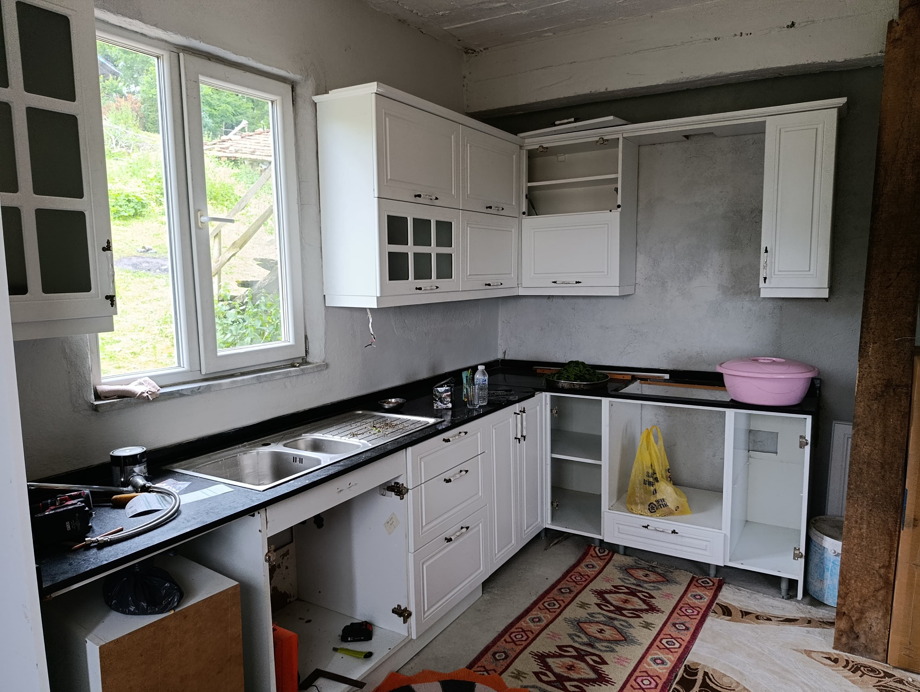 An L-shaped modular kitchen cabinet system, presented in an aerial view. It features white shaker-style cabinet doors and drawers for both upper and lower units. The lower cabinetry includes an integrated double-basin stainless steel sink within a black speckled countertop. One section of the upper cabinets features glass-front doors, providing a display option. All cabinets are equipped with simple silver-toned handles. A light grey backsplash area is visible between the upper and lower units, against a white background. — 3D model