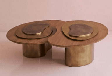 A contemporary coffee table featuring a biomorphic design with a dark wooden top and antique brass-toned metal elements. The tabletop consists of two conjoined organic shapes, each with nested brass-toned inlays. The table is supported by two substantial, organically shaped brass-toned cylindrical bases. — 3D model