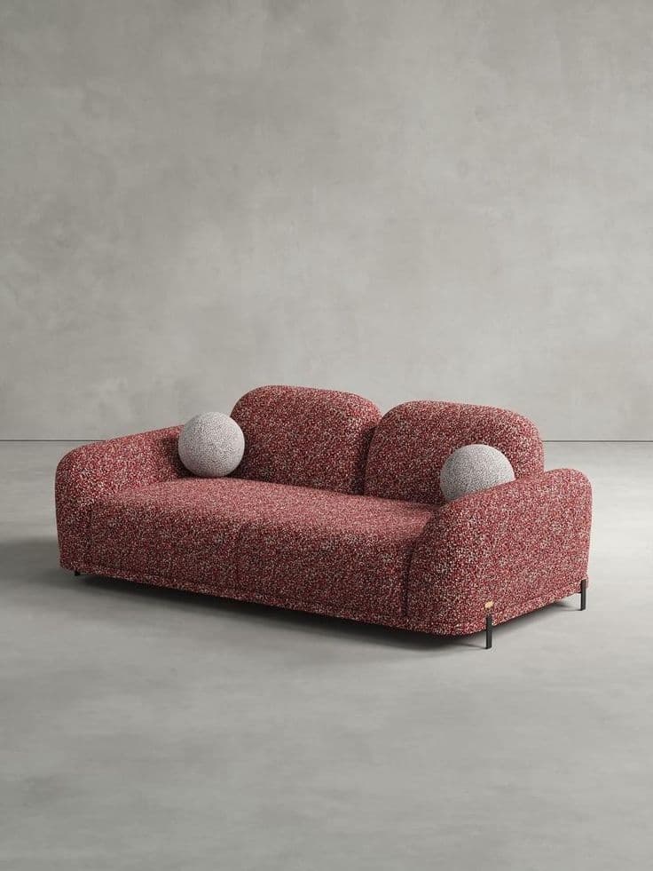 A modern two-seater loveseat featuring a distinctive speckled red and white textured upholstery, likely boucle or a similar fabric. The loveseat exhibits soft, rounded edges and voluminous forms across its armrests, seat, and two separate back cushions. It is supported by thin, minimalist black metal legs accented with brass tips. Two spherical throw pillows in a lighter speckled fabric are positioned on the sofa. — 3D model