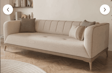 A contemporary three-seater sofa upholstered in a light beige fabric, featuring vertical channel tufting on the backrest and button tufting on the seat cushion. It has sloped, rounded arms and rests on tapered wooden legs with a visible wooden trim along the bottom edge of the frame. The sofa includes several decorative throw pillows in coordinating neutral tones. — 3D model