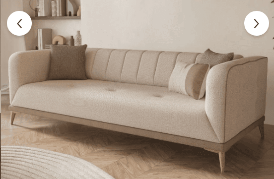 A contemporary three-seater sofa upholstered in a light beige fabric, featuring vertical channel tufting on the backrest and button tufting on the seat cushion. It has sloped, rounded arms and rests on tapered wooden legs with a visible wooden trim along the bottom edge of the frame. The sofa includes several decorative throw pillows in coordinating neutral tones. — 3D model