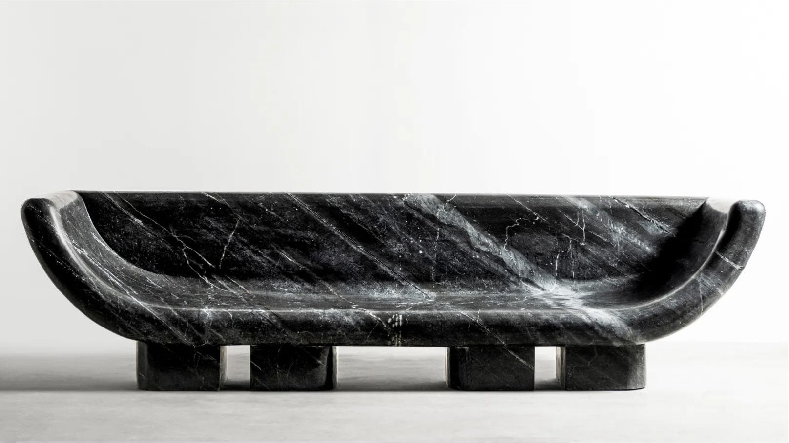A monolithic daybed or bench carved from black marble with prominent white veining. It features a scooped, ergonomic form that curves upwards at both ends, serving as integrated armrests and backrest. The substantial form is supported by two pairs of thick, rectangular block legs, creating a robust and sculptural appearance. — 3D model