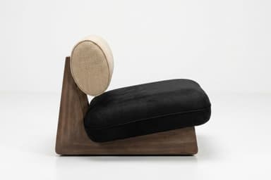 A modern, low-profile lounge chair featuring a distinctive solid wood base. The base is dark-toned wood, shaped with a geometric, wedge-like profile. It supports a plush, rectangular black seat cushion, likely upholstered in velvet or microsuede. The backrest is a cylindrical bolster cushion, upholstered in a textured light beige fabric, resting against the angled wooden support structure. — 3D model