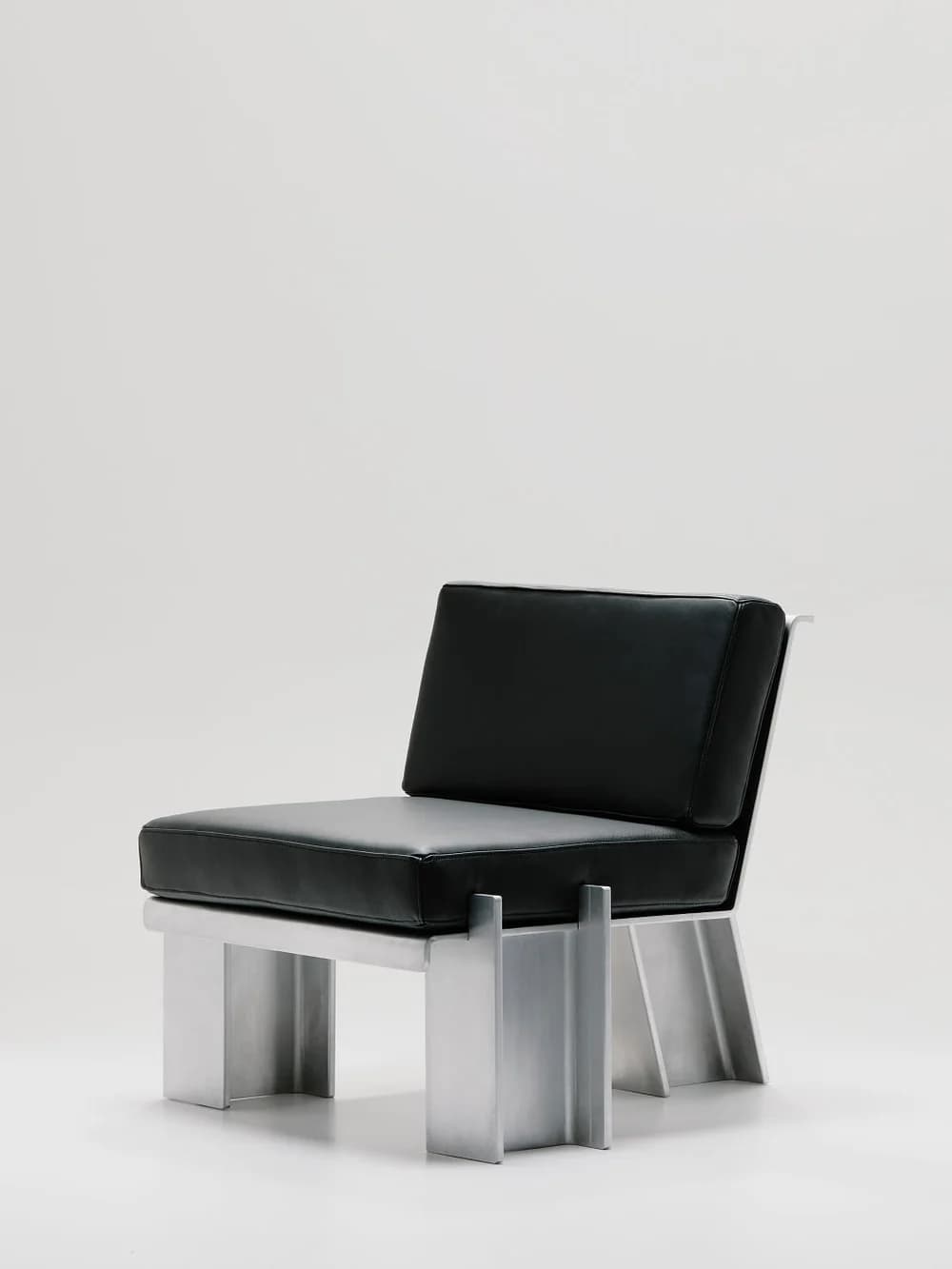 An armless lounge chair featuring a robust, angular frame constructed from brushed or polished aluminum, supporting thick, box-style cushions upholstered in black leather. The design is notably minimalist and industrial, characterized by its geometric lines and solid, block-like legs formed from metal channels. The seat and back cushions provide a clear visual contrast against the metallic frame. — 3D model