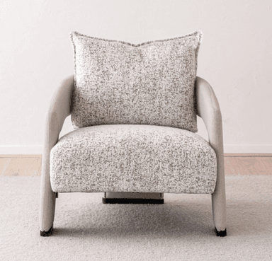 A contemporary upholstered armchair featuring a rounded silhouette and a neutral-toned, speckled fabric. The chair includes integrated, curved armrests and an upholstered backrest, complemented by a separate, plush back cushion with piped edges. The seat appears integrated, and the chair stands on short, upholstered legs or a low, concealed base, suitable for modern interior settings. — 3D model