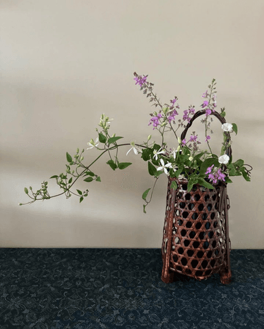 A traditional Japanese ikebana vase, meticulously crafted from dark, lacquered woven bamboo in a hexagonal pattern. It features a footed base for stability and an elegant arched handle. The vase holds a floral arrangement with delicate purple and white blossoms and trailing green foliage, presented against a stark white background. — 3D model