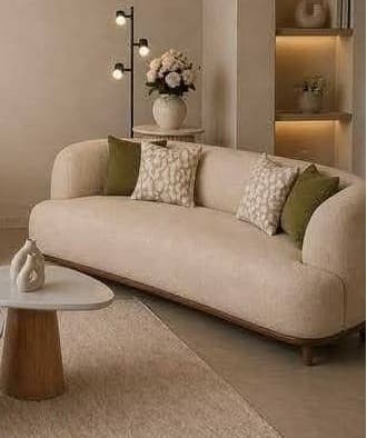 A contemporary curved sofa upholstered in light beige fabric, featuring a continuous rounded backrest and arms. The sofa sits on a recessed dark wood plinth base with short, tapered wooden legs. It is accessorized with four decorative pillows, two solid olive green and two with a patterned beige and cream design. — 3D model