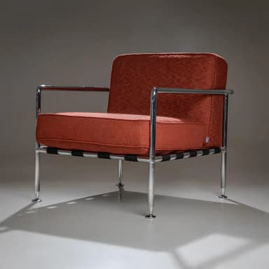 A pair of armchairs with a modern design. Each chair features a polished chrome frame, including the armrests and legs. The seating and back cushions are upholstered in a textured, rust-colored fabric. The seat support appears to be constructed from woven straps. The chairs have a minimalist aesthetic, emphasizing clean lines and industrial materials. — 3D model