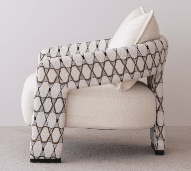 A contemporary upholstered armchair featuring a plush, light-colored bouclé fabric on the seat and back, complemented by patterned armrests and legs with a geometric design in black, white, and a coppery tone. It includes a matching back cushion. — 3D model