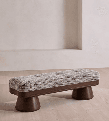 A contemporary rectangular bench featuring a deeply cushioned, tufted upholstered top. The upholstery is a textured, multi-tonal fabric in shades of brown, gray, and white. The base consists of a dark-stained wooden frame from which two substantial, turned wooden pedestal legs descend, providing stable support. The design combines comfort with a modern aesthetic, highlighted by the striking silhouette of the legs. — 3D model