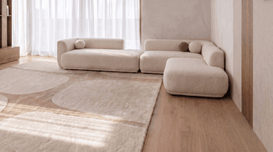 A modular sectional sofa with a low profile and rounded armrests. The sofa is upholstered in a textured light-colored fabric and features a configuration of multiple seating units that can be rearranged. It is accompanied by several decorative throw pillows and a draped throw blanket. The overall aesthetic is modern and minimalist. — 3D model