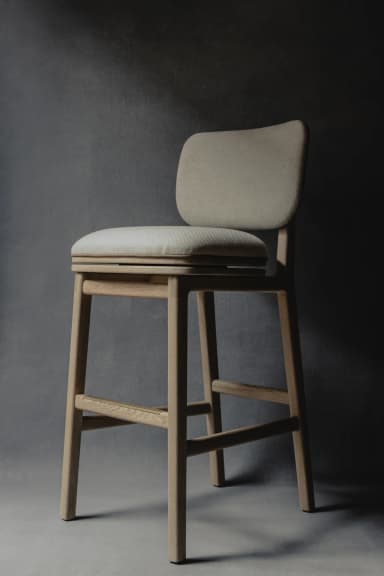A close-up view of the back and side of a modern dining chair. The chair features a light-toned wooden frame, possibly ash or beech, with visible joinery. The backrest and seat are upholstered in a textured, light beige or cream fabric. A distinctive design element is a horizontal strap of light tan leather, secured around the backrest frame, adding a touch of contrast and craftsmanship. The cushioning appears ample and comfortable. — 3D model