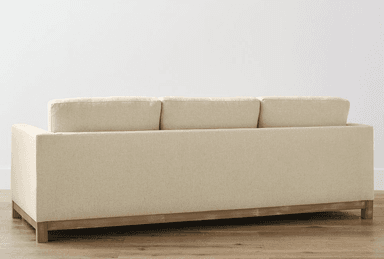 A three-seater sofa upholstered in a textured, light beige fabric. The sofa features a wooden base frame and legs, with a visible grain texture on the wood. The back of the sofa is designed with three separate cushions, and the side profile shows a clean, rectangular form. The overall aesthetic suggests a contemporary or transitional style. — 3D model