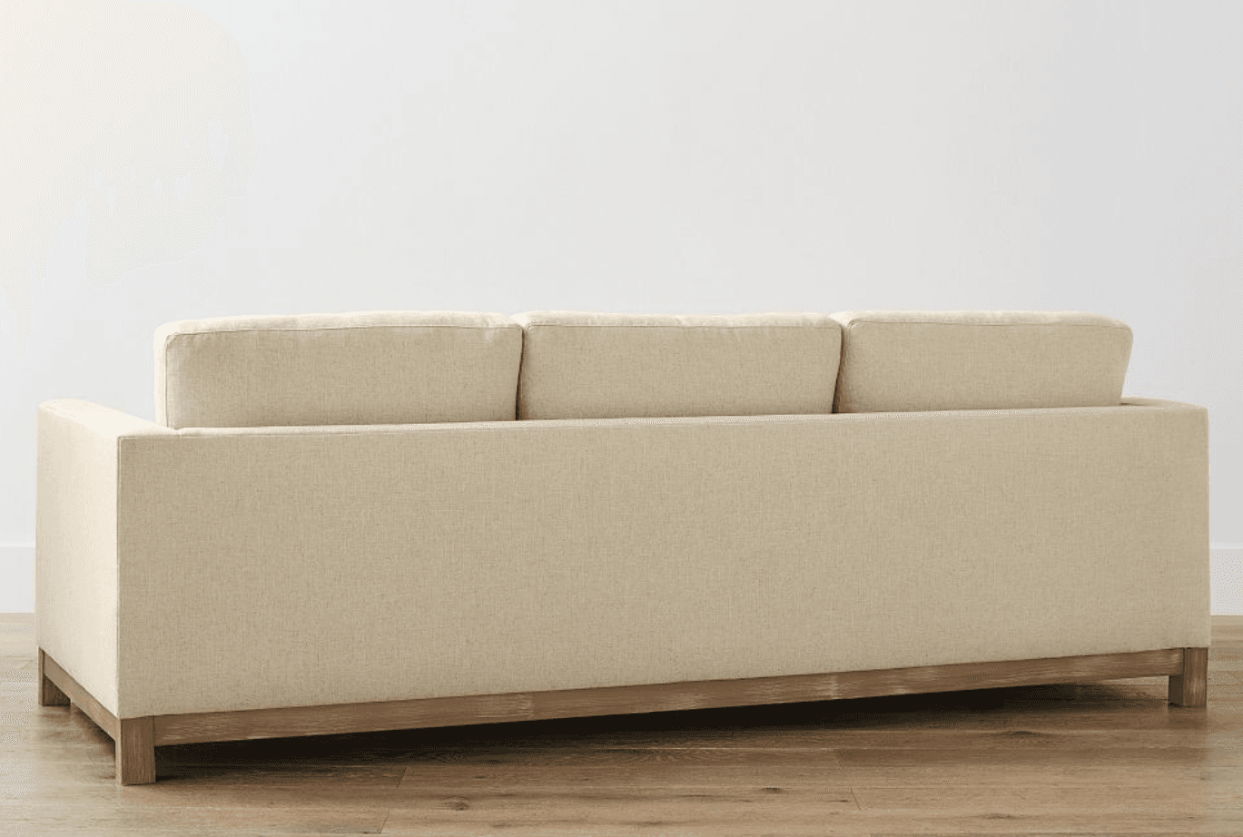 A three-seater sofa upholstered in a textured, light beige fabric. The sofa features a wooden base frame and legs, with a visible grain texture on the wood. The back of the sofa is designed with three separate cushions, and the side profile shows a clean, rectangular form. The overall aesthetic suggests a contemporary or transitional style. — 3D model