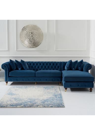 A luxurious blue velvet Chesterfield-style sectional sofa with a right-hand facing chaise lounge. It features deep button tufting across the backrest and inner arms, classic rolled arms, and antique brass nailhead trim along the base and arm fronts. The sofa stands on dark wood bun feet with brass caster caps, contributing to its elegant and traditional aesthetic. — 3D model