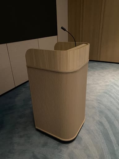 A modern lectern, also known as a podium, constructed from light-toned wood. It features a curved top surface designed to hold notes or a book for a speaker. The body of the lectern is characterized by vertical fluting, adding textural interest and a contemporary aesthetic. It stands on a recessed black plinth base, providing stability and a subtle contrast to the light wood. — 3D model