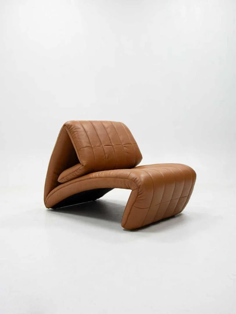 A unique contemporary lounge chair featuring a distinctive folded form upholstered in quilted brown leather. The chair has a continuous, sinuous profile with a padded seat and backrest, emphasizing comfort and modern aesthetics. Its design suggests a low-slung, relaxed posture. — 3D model