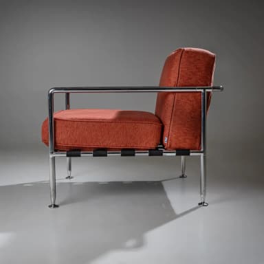 A pair of armchairs with a modern design. Each chair features a polished chrome frame, including the armrests and legs. The seating and back cushions are upholstered in a textured, rust-colored fabric. The seat support appears to be constructed from woven straps. The chairs have a minimalist aesthetic, emphasizing clean lines and industrial materials. — 3D model