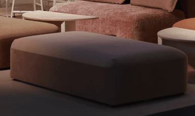 A rectangular upholstered ottoman featuring soft, muted pink fabric and a contemporary design with gently rounded corners. The piece exhibits a seamless appearance with a subtle seam line delineating the top from the side panels, giving it a solid, block-like form. — 3D model