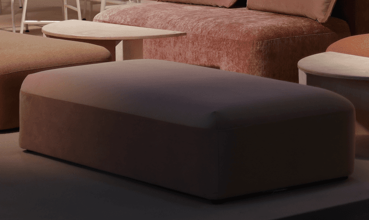 A rectangular upholstered ottoman featuring soft, muted pink fabric and a contemporary design with gently rounded corners. The piece exhibits a seamless appearance with a subtle seam line delineating the top from the side panels, giving it a solid, block-like form. — 3D model