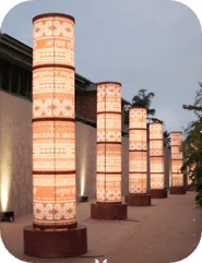 A tall, cylindrical floor lamp featuring a segmented body composed of multiple stacked sections, each adorned with intricate orange and white patterns including script. It has a round, dark wooden base and a matching top. — 3D model