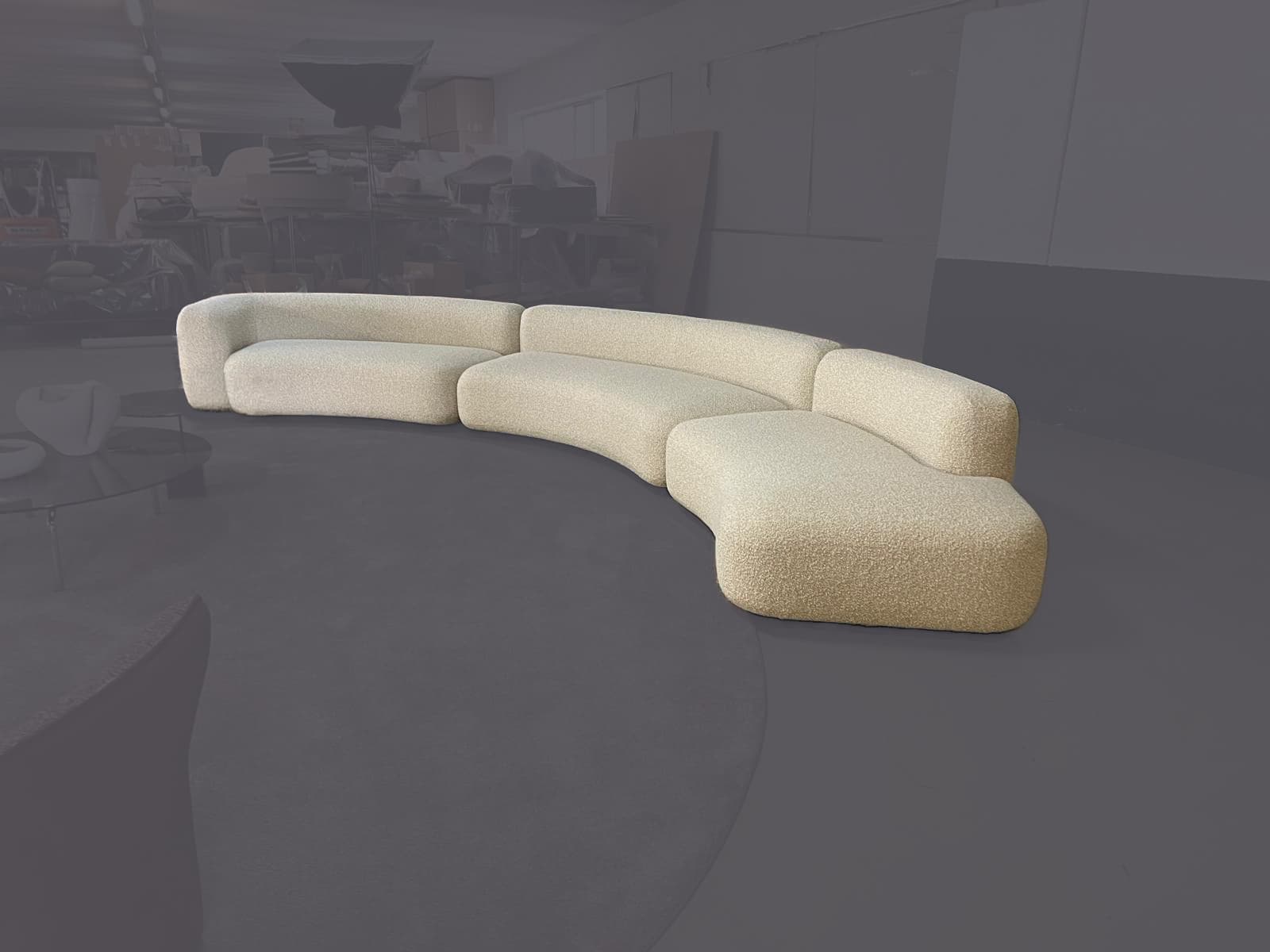 A large, modular sectional sofa with a distinct curved or serpentine silhouette, upholstered in a textured, light-colored boucle fabric. The sofa consists of multiple individual sections that can be configured to create various arrangements. It features a low-profile design with rounded edges and continuous, softly cushioned forms. — 3D model