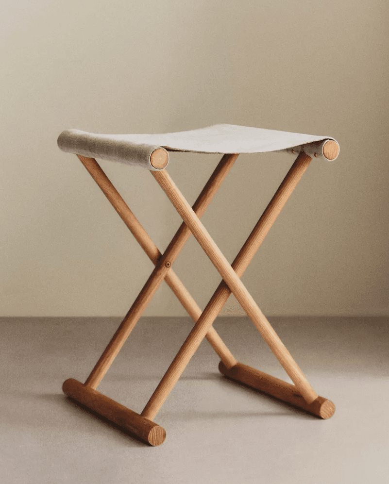 A lightweight, portable folding stool featuring an X-frame design crafted from light-toned wood. The seat is a piece of beige or off-white canvas fabric stretched between the top wooden bars, creating a comfortable sling-like surface. The construction allows the stool to collapse for easy storage and transport. — 3D model