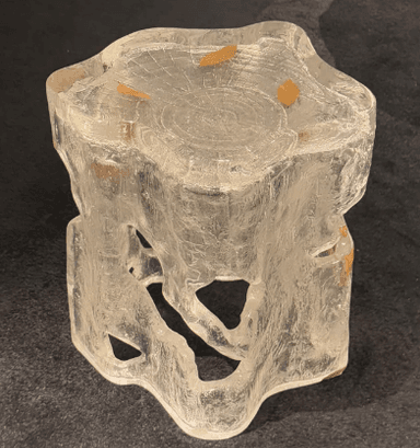 A sculptural accent table or stool crafted from clear, textured resin, designed to resemble a block of ice or a natural tree stump. The piece features an organic, irregular silhouette with cut-out sections in its body, giving it a light, ethereal appearance. Embedded within the clear material are small, delicate golden leaf inclusions. The top surface is flat but textured with an embossed wood grain pattern, suggesting tree rings, further enhancing its nature-inspired aesthetic. The overall impression is one of modern artistry combined with natural elements. — 3D model