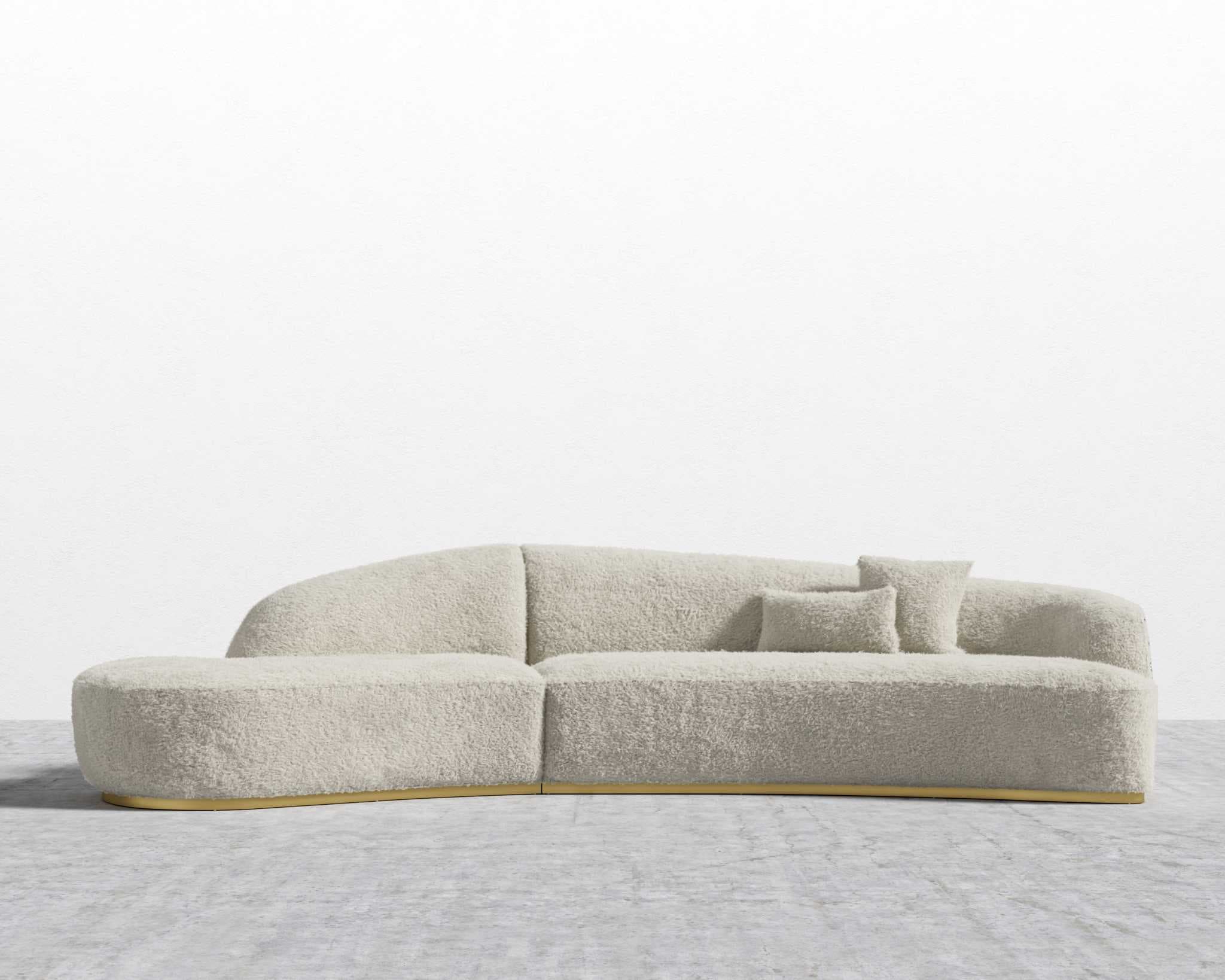 A contemporary modular sectional sofa featuring an asymmetrical, curved design. It is upholstered in a light beige or cream-colored boucle fabric, providing a textured, soft appearance. The sofa sits on a low-profile golden or brass-toned base, which accents its modern aesthetic. The configuration includes at least two modules, one with a rounded end, and is accompanied by two small, matching toss pillows. — 3D model