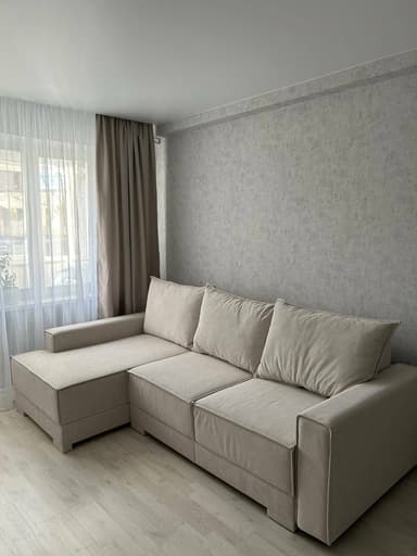 An L-shaped sectional sofa upholstered in a light beige fabric, featuring an integrated left-hand facing chaise lounge. The sofa has a clean, modern aesthetic with visible stitching details, low-profile block feet, and three separate back cushions. The armrests are broad and flat, matching the overall minimalist silhouette. — 3D model