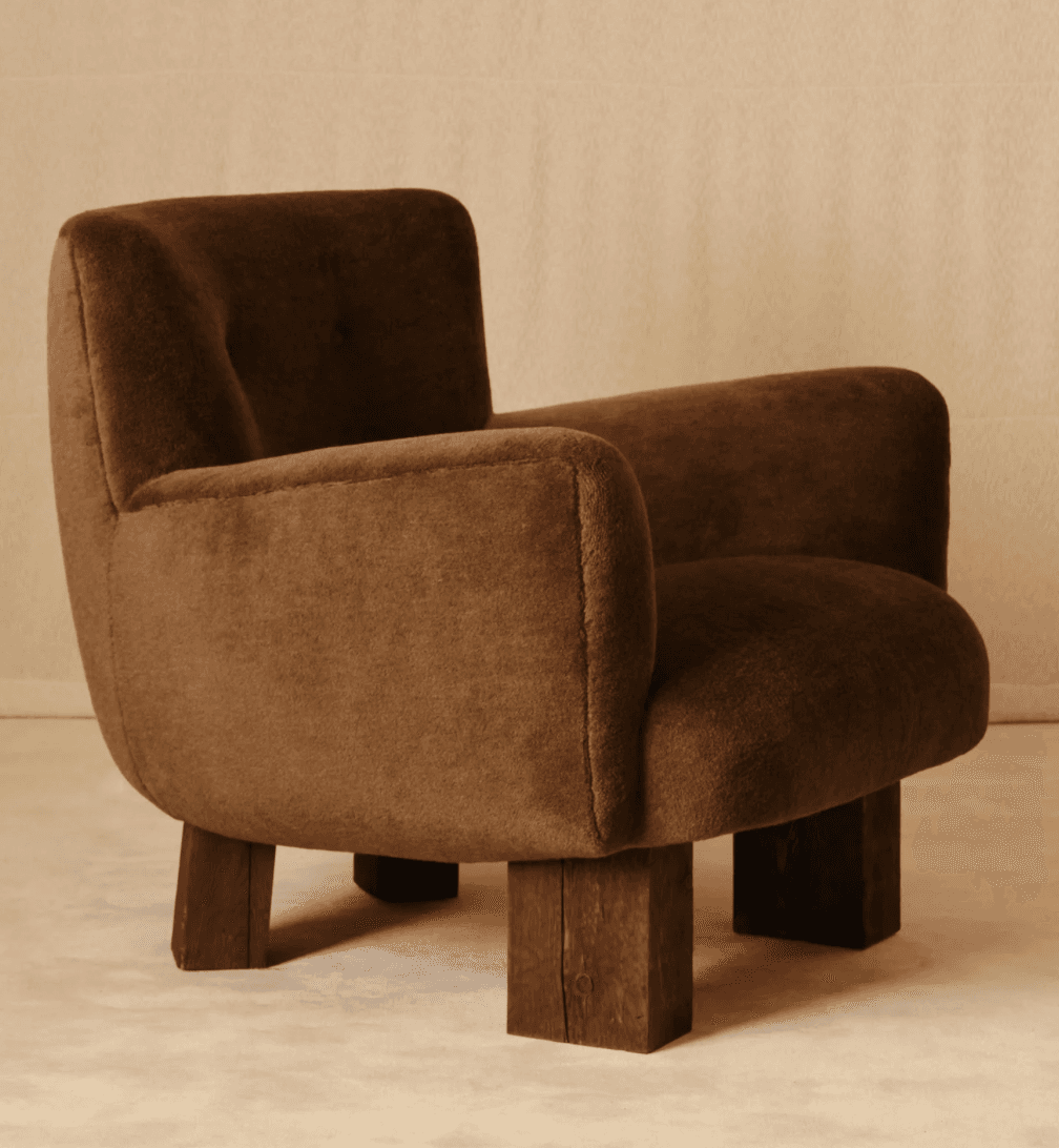 A plush, upholstered armchair with rounded edges and broad armrests. The chair is supported by four thick, dark wooden block legs that provide a stark contrast to the soft upholstery. The upholstery appears to be a textured fabric, possibly velvet or a similar material. — 3D model
