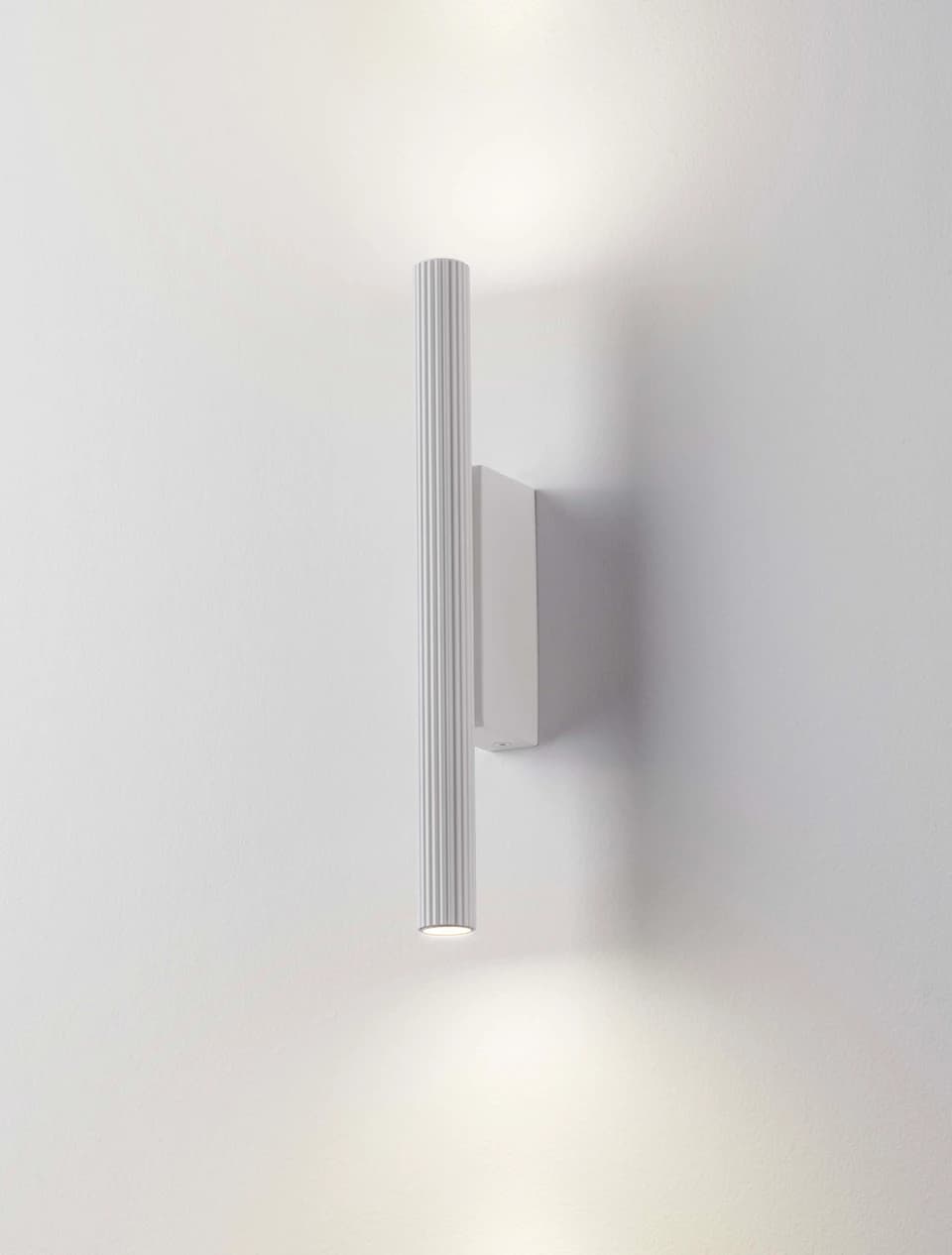 A contemporary white wall sconce featuring a vertical, cylindrical body with a distinctive ribbed texture. The fixture is mounted directly to the wall and emits light from both its upper and lower ends, providing an up-and-down illumination effect. — 3D model