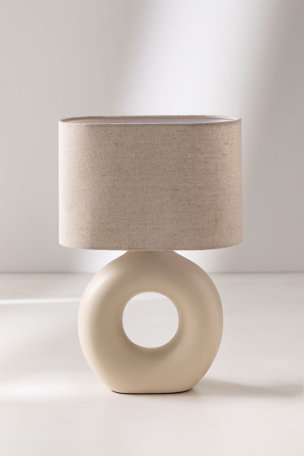 A modern table lamp featuring a cream-colored ceramic torus-shaped base with a central cut-out, topped with a cylindrical light beige fabric shade. — 3D model