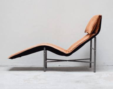 A modern chaise lounge with a thin, tubular metal frame supporting a flexible, reclining seat and back. The upholstery appears to be a tan-colored, possibly leather or suede, material with visible stitching and tufting in the seat area. A separate headrest cushion is positioned at the top of the chaise. The frame has a matte silver or grey finish. — 3D model