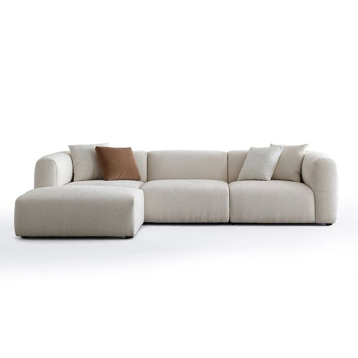 A modern modular sectional sofa upholstered in light beige textured fabric, featuring a low-profile design with plump, rounded cushions and broad arms. The configuration includes a left-hand chaise extension and two additional armless seating segments, forming an L-shape. Three decorative throw pillows are visible: one solid brown and two lighter, subtly patterned ones. — 3D model