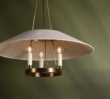 A modern chandelier featuring a wide, shallow, inverted dome-shaped lampshade made of light gray fabric. Beneath the fabric shade, a polished brass ring holds three vertically oriented light sockets, each fitted with a candle-style LED bulb. The fixture is suspended by thin brass rods, contributing to its minimalist and elegant aesthetic. The light emitted by the bulbs is diffused by the fabric shade, creating a soft, ambient glow. — 3D model